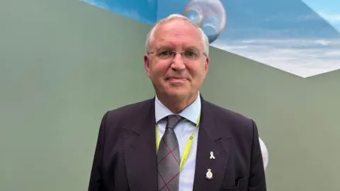 Chris Nelson, an older man with thin framed glasses, who is wearing a black blazer, a grey and red tie, and a light-coloured shirt, smiles as he stands in front of a green and blue background. There are two enamel pins on his lapel.