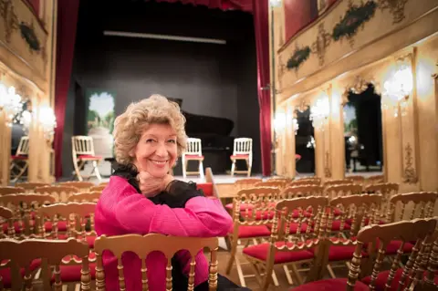 A woman with short curly hair dressed in black and pink leans on the back of a chair and smiles to a camera while sitting in a theatre space.