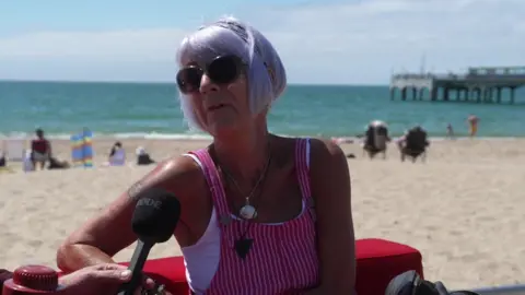 Denise is wearing a pink and white striped pinafore top and is sitting on a red sofa in front of the beach