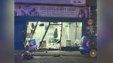 Smashed-up window at the beauty salon where the crash happened. Glass and rubble lies in the road. It is dark but lights are still working in the damaged building.