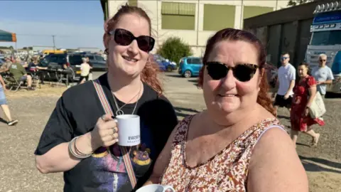 Phillip Norton/BBC. Aimee y Lynn Ledger posan con sus tazas. Aimee lleva una camiseta negra con un colorido estampado en la parte delantera. Lynn lleva un vestido marrón con un estampado de flores blancas.