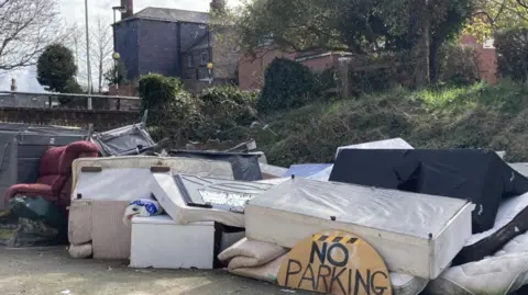 A pile of mattresses, beds and other furniture lying in the street with a wooden no parking sign in front