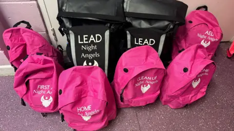 Chrissie Lowery A pile of pink backpacks printed with the Night Angels logo in white. Each one is labelled to match their respective roles and purposes, including first aid, AED and clean up. Two larger black backpacks are also among the pile - they are also printed with the Night Angels logo and labelled with the word lead in capital letters. 