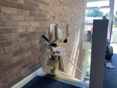 A white stairlift at the top of a flight of stairs. It is folded up against a brick wall.