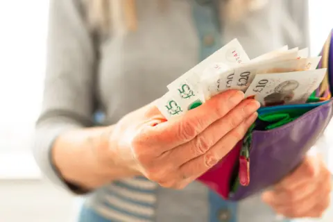 A hand grasps £5, £20 and £10 notes protruding from a purple purse