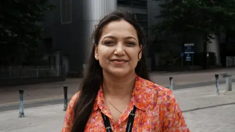John Fairhall/BBC Shabnam Sagar wears an orange shirt with floral pattern, with a dark lanyard around her neck. She has long dark hair past her shoulders. She stands in front of a dark building and is smiling at the camera.