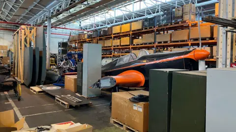 An old black and orange lightweight aircraft on the floor of a huge warehouse. It is surrounded by large RAF objects with shelves containing many boxes behind it.