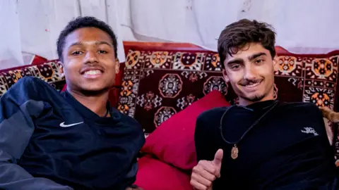 BBC/Studio Lambert Jo and Kush smile as they lean back on maroon patterned cushions in a Turkish home.