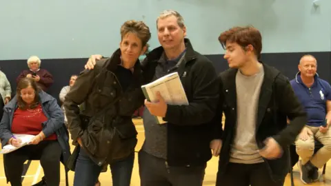 BBC Deputy Heidi Soulsby, who is wearing a green Barbour jacket being hugged by her husband Phillip. Their son is standing next to them. 