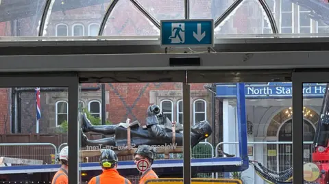BBC A statue of potter Josiah Wedgwood, lay on its back, is being transported past the frontage of a railway station. It is viewed through a set of glass windows.
