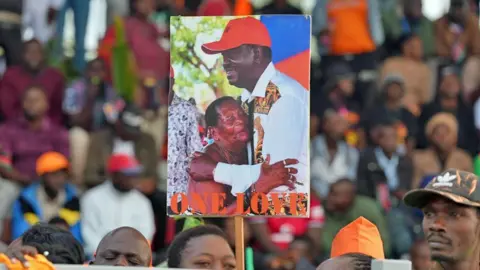 A female mourner in a crowd at a stadium in Kisumu holds up a poster showing Raila Odinga hugging a woman - underneath in red are the words 'One Love'.