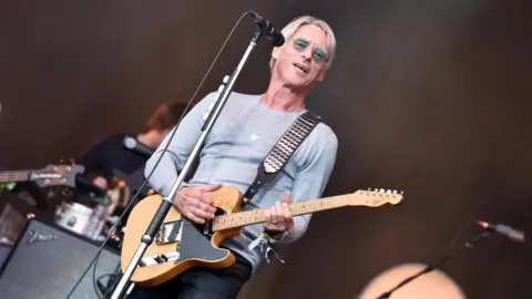 BBC / Steve Barney Paul Weller, performing with a guitar and a microphone he is smiling out off stage and in the background other musicians can be seen.