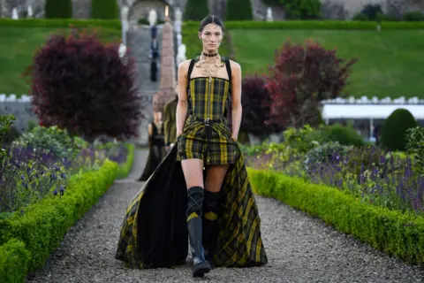 Getty Images A dark-haired model walks the runway - a long path through the beautiful gardens of Drummond castle. She wears a modern outfit in yellow and black tartan - with a fitted corset top and matching shorts and long socks, with a long tartan train. She wears a striking piece of intricate costume jewellery round her neck. other models descend steep steps behind her.