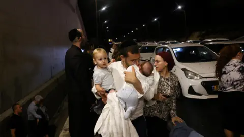 Reuters A man holds children as people take cover as air raid sirens wail across Israel during an Iranian missile attack in central Israel (October 1, 2024)