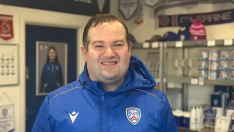 Jonathan Fillis is a man with dark short hair. He is standing in the shop with cups and mugs on shelves behind him, smiling at the camera.
He is wearing a bright blue sports coat with the Coleraine FC emblem on the right hand side.