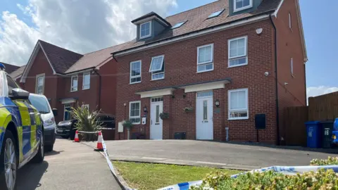 Ian Brown/BBC A pair of semi-detached red brick houses, that are both three storeys high. They have white windows and doors. A police car can be seen in the left-hand corner. It is sunny.