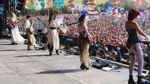 A line of five women in various costumes, ranging from flowing white dress to a black basque. They are singing on a large stage, three have guitars and one is playing a keyboard. There is a huge crowd beyond the stage, and flags and balloons can be seen above them.