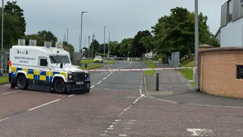 A police vehicle is beside a police cordon.
Another police car is stationed further up a road beside another cordon.
