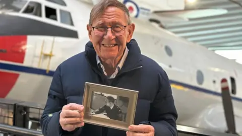 A man wearing a blue coat and shirt and glasses and smiling at the camera. He is holding a black and white photo of himself as a young man wearing an RAF uniform. 