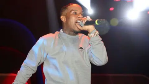 Getty Images Tory Lanez se apresenta no palco. Ele está vestindo um suéter cinza e cantando no microfone. Ele fica na frente de um fundo preto. 