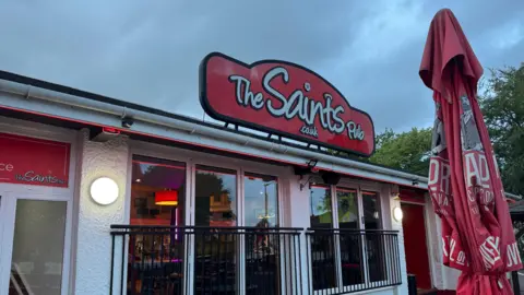 Exterior of the pub with large glass windows and a red sign on top in Southampton FC colours.
