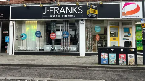 BBC/Charlotte Rose A shop window with black heading and white writing saying "J Franks Bespoke Interiors". The shop window still has samples of curtains and shutters, and there are stickers with sales and offers.