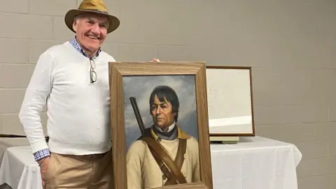 D'Arcy Jenish A middle aged man wearing a cream jumper and a hat, standing next to a portrait of David Thompson. D'Arcy Jenish is one of the leading authorities on David Thompson