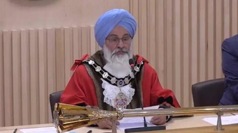 A man wearing a blue turban wearing official mayoral dress sitting at a brown desk with a gold coloured stick in front of him on the desk.