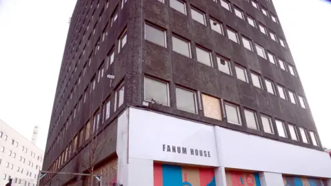 A grey tower block with some of its windows boarded up. On the ground floor a sign that reads "FANUM HOUSE"