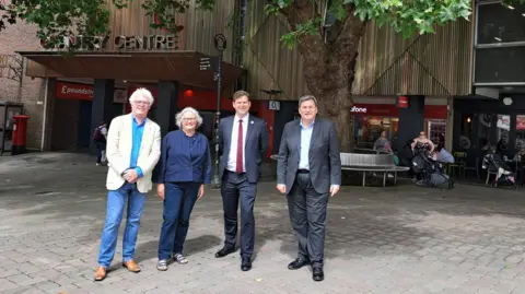 ThinkAndoverTownCentre From left to right Aidan Ridyard and Helen Grassly of BFF, councillor Phil North and councillor Kit Malthouse MP, outside of the location for the new theatre in Andover. They are smiling at the camera. The Chantry Centre is in the background. A woman with a pram appears to be speaking to another woman who is sitting on a round metal bench. A man is passing by a shop.   