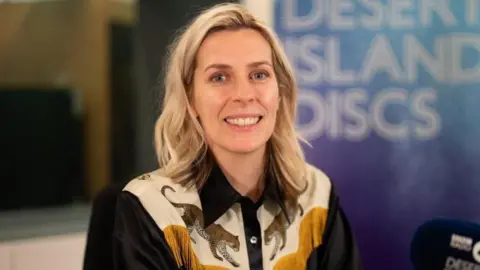 BBC/PA Sara Pascoe, who has shoulder-length blonde hair and wears a fringed black shirt with leopards on it, smiles as she sits in the Desert Island Discs studio