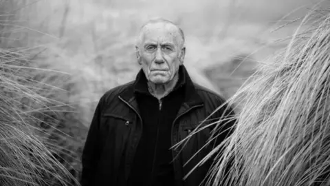 Stu McKenzie Black and white picture of Richard Lee. He is dressed in a black sweater and jacket between tufts of long grass in sand dune