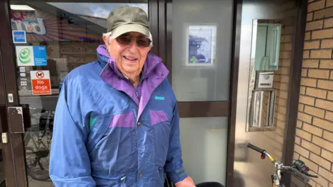 BBC/Elliott Green An elderly man is wearing a khaki coloured baseball cap, a brightly coloured blue and purple rain coat and sunglasses. He is stood by a door holding a bike