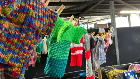BBC/Cathy Killick A washing line in a barn with a number of colourful small jumpers pegged on it.