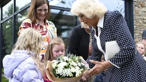 PA Media Queen Camilla wearing a black and white polka dot dress with a white collar received a bouquet of white flowers from two young girls. A woman standing behind the girls looks at them smiling.
