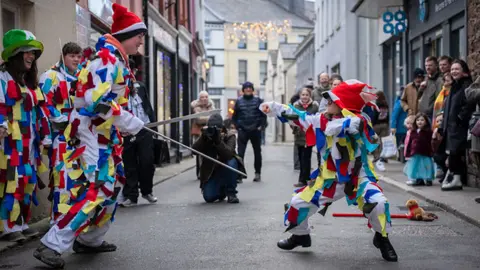 Brook Wassall Two people dressed in white uniforms decorated with colourful paper look at each other - one is holding sword props. Onlookers gather on the road and pavements, with shop signs and Christmas lights in view.
