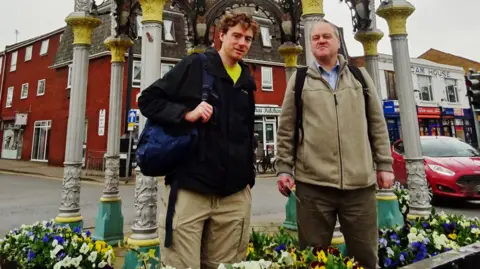 Colin Bell Two men are stood in a flower bed while they pose for a photo in a town centre