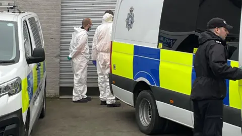 PA Media Part of the side of a police car can be seen on the left hand side and the side of a police van on the right hand side. They are parked outside a white brick building with a white metal roll-down door visible. A police officer dressed in black is standing by the van on the right. Two forensic officers in white suits are standing by the roll-down door behind the vans with their backs to the camera.