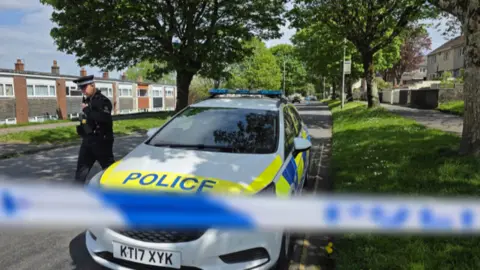 The image shows a residential street closed off by blue-and-white police tape, with a marked police car parked at the roadside under trees. A uniformed police officer is standing beside the vehicle, while low-rise homes and grass verges line the quiet road in the background.