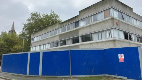 An abandoned office office block with rows of glass windows and grey bricks, stained green by moss. It is surrounded by high blue panels, and has trees and the spire of Truro Cathedral in the background.