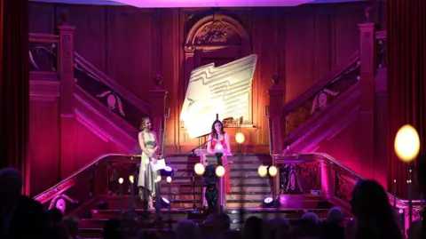 Press Eye Two women, one speaking at a podium and both wearing evening dresses, on stage at the awards ceremony. The stage is below two staircases and is lit in purple lighting