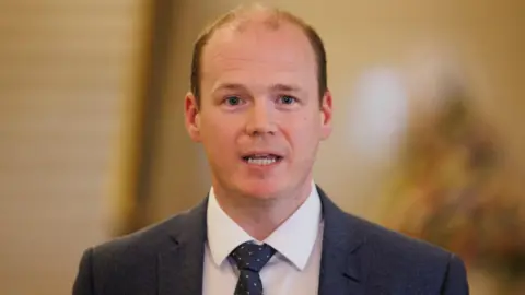 PA Media Communities Minister Gordon Lyons pictured during a press conference at Parliament Buildings at Stormont in Belfast. He has thinning brown hair. He is wearing a dark suit, white shirt and navy tie with pink spots.