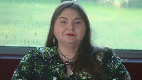 A woman with long dark hair and dark eyes is looking directly at the camera. She is sitting in front of a rain covered window, with grass visible behind her. The woman, who is wearing red lipstick and long ear rings, is dressed in a green patterned top and has a decorative necklace on. 