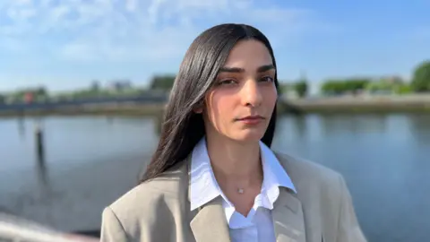 Ellie Wilson, who has long, straight dark hair, poses on the Millennium Bridge in Glasgow wearing a white shirt and a beige blazer.