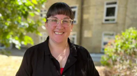 She is wearing a red top paired with an oversized black cardigan. Her dark brown hair is in a fringe and she is is wearing clear framed glasses, highlighted in gold.
 She has one hand on her hip.
