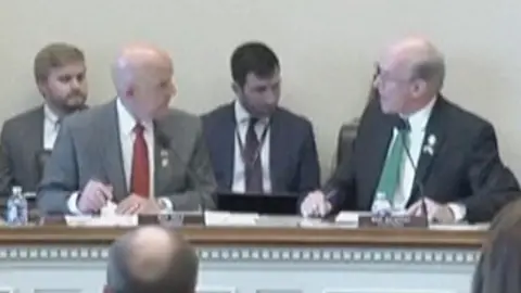 A house hearing room is seen from the back with lawmakers on a dais and witnesses facing them.