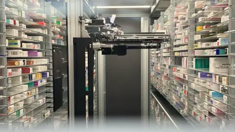 A black robotic device is positioned in the middle of a room. On either side are shelves stacked with various types of medication. There is a dark grey door at the far end of the room behind the robot.