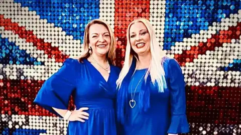 Supplied Two women wearing blue dresses standing in front of a union jack backdrop