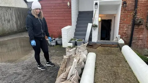 BBC/George Carden A woman stands next to a footpath with sandbags either side. The path leads to a basement flat. There is a large puddle behind her.