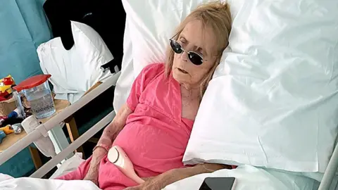 Jackie is lying in a hospital bed, wearing a pink gown. She is wearing a pair of sunglasses and looks unwell. 